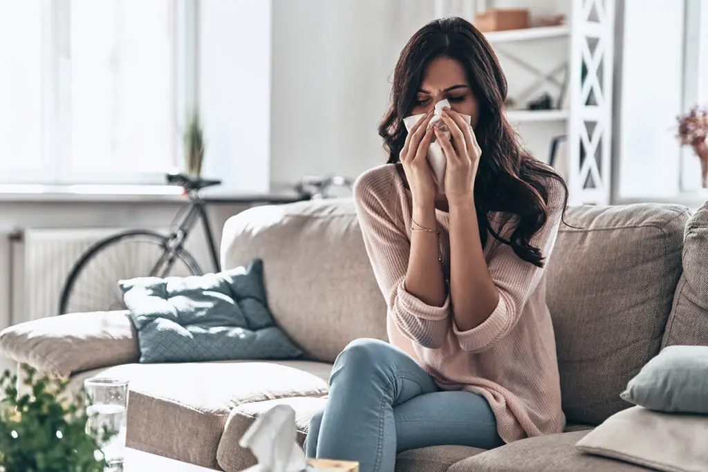 Cold and flu. Sick young woman blowing the nose using tissue paper while sitting on the sofa at home, household allergies